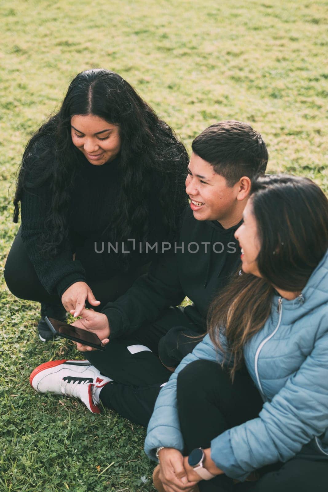 Hispanic male teenager holding smartphone looking away while sitting on grass with Hispanic mother and sister in park on sunny day by carlosviv