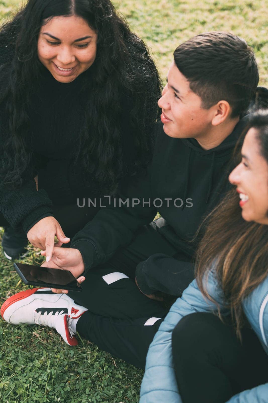 three people of hispanic-latino ethnicity sitting on the green grass of a park, with smart device by carlosviv