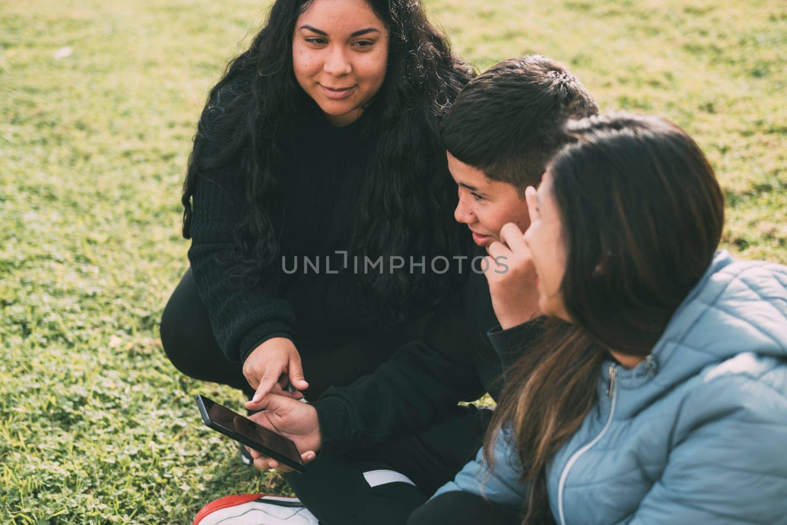 Hispanic family spending quality time together in a local park on a beautiful, sunny day. A teenage boy is sitting on the grass, holding his smartphone while looking away. His mother and sister are sitting next to him, looking at the camera and smiling