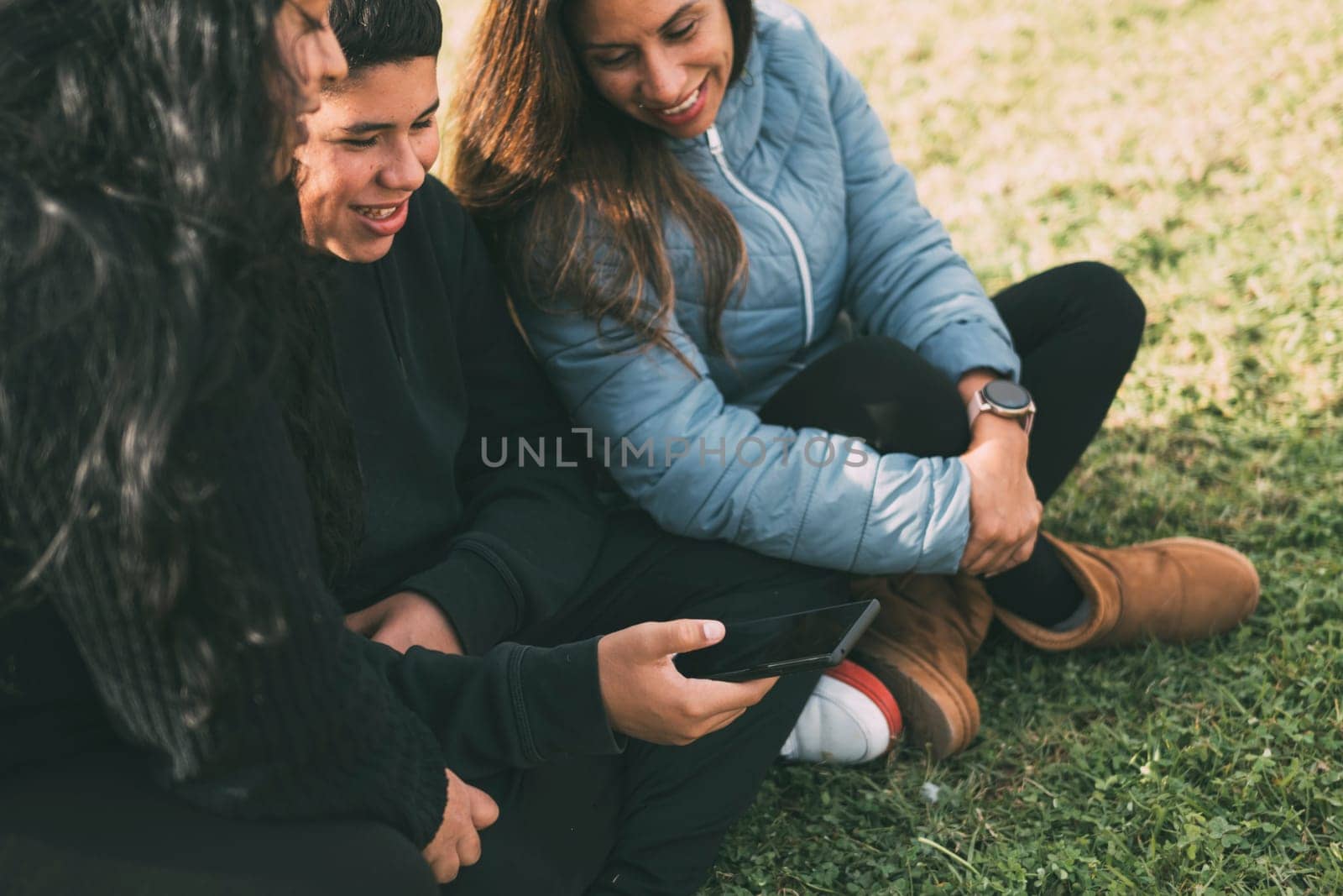 Hispanic family spending quality time together in a local park on a beautiful, sunny day. A teenage boy is sitting on the grass, holding his smartphone while looking away. His mother and sister are sitting next to him, looking at the camera and smiling.