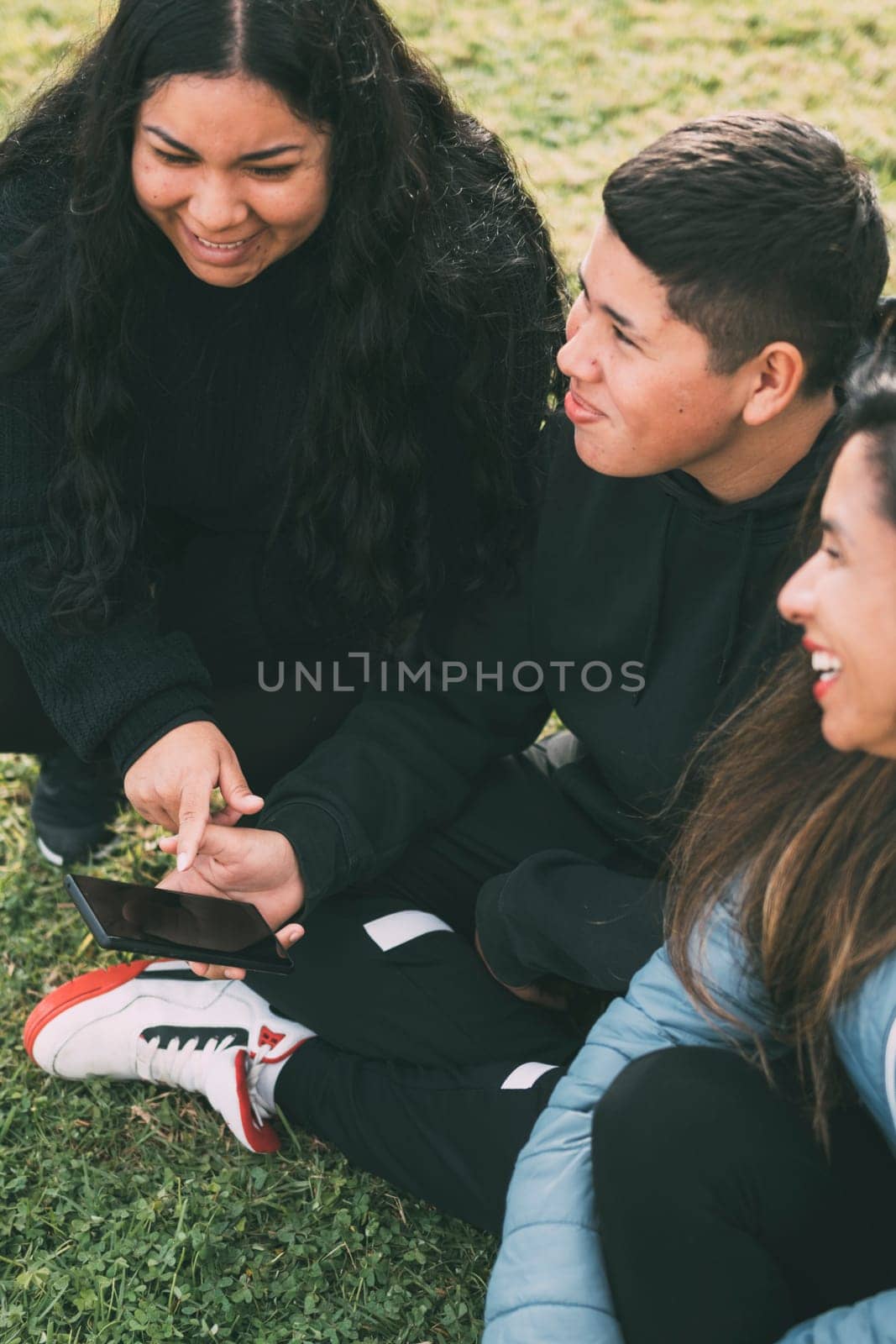 three people of hispanic-latino ethnicity sitting on the green grass of a park, with smart device. relaxed and comfortable as they entertain themselves with their screens, browsing social networks, dressed in casual clothes and their faces suggest satisfaction with their activity. it conveys a sense of modernity and connectivity that is common in today's digital age.