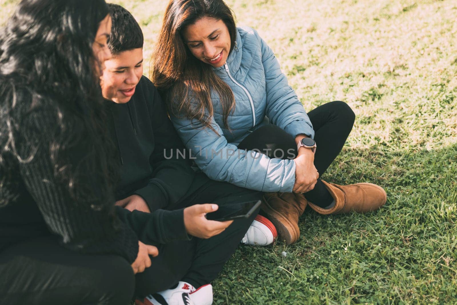 Hispanic family spending quality time together in a local park on a beautiful, sunny day. A teenage boy is sitting on the grass, holding his smartphone while looking away. His mother and sister are sitting next to him, looking at the camera and smiling