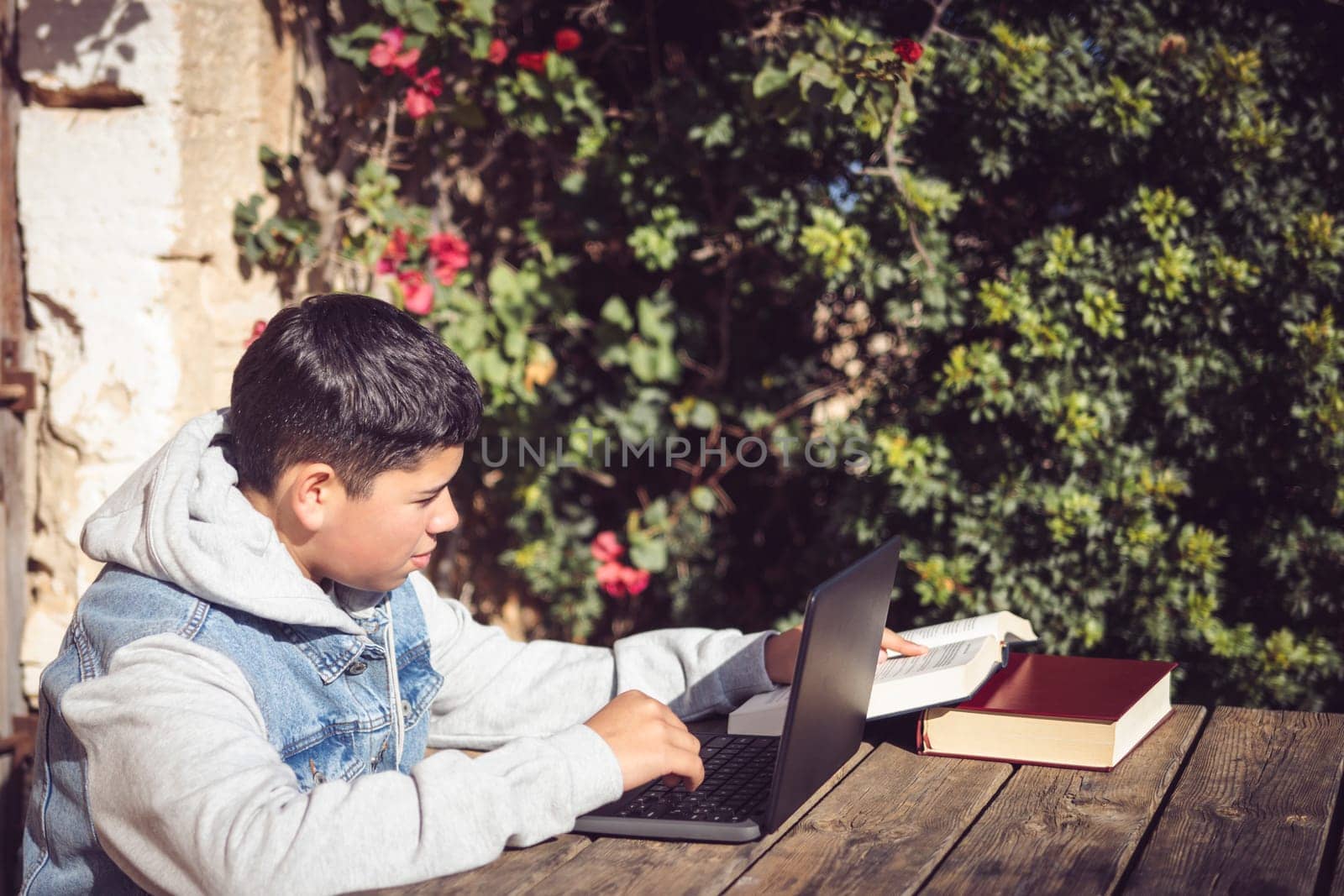 young latin man reads book in front of laptop, outdoors in city park, doing high school study work