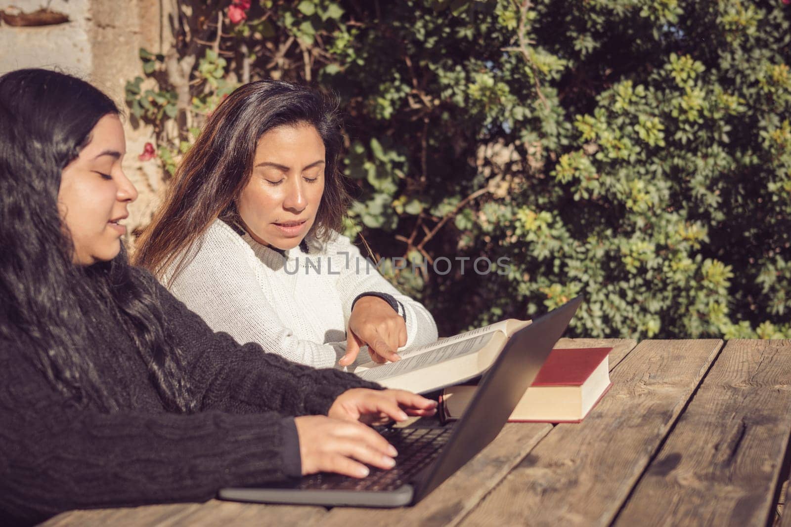 two latin women with long dark hair, working outdoors in the park with books and laptops, sitting at a wooden table with a natural green background.