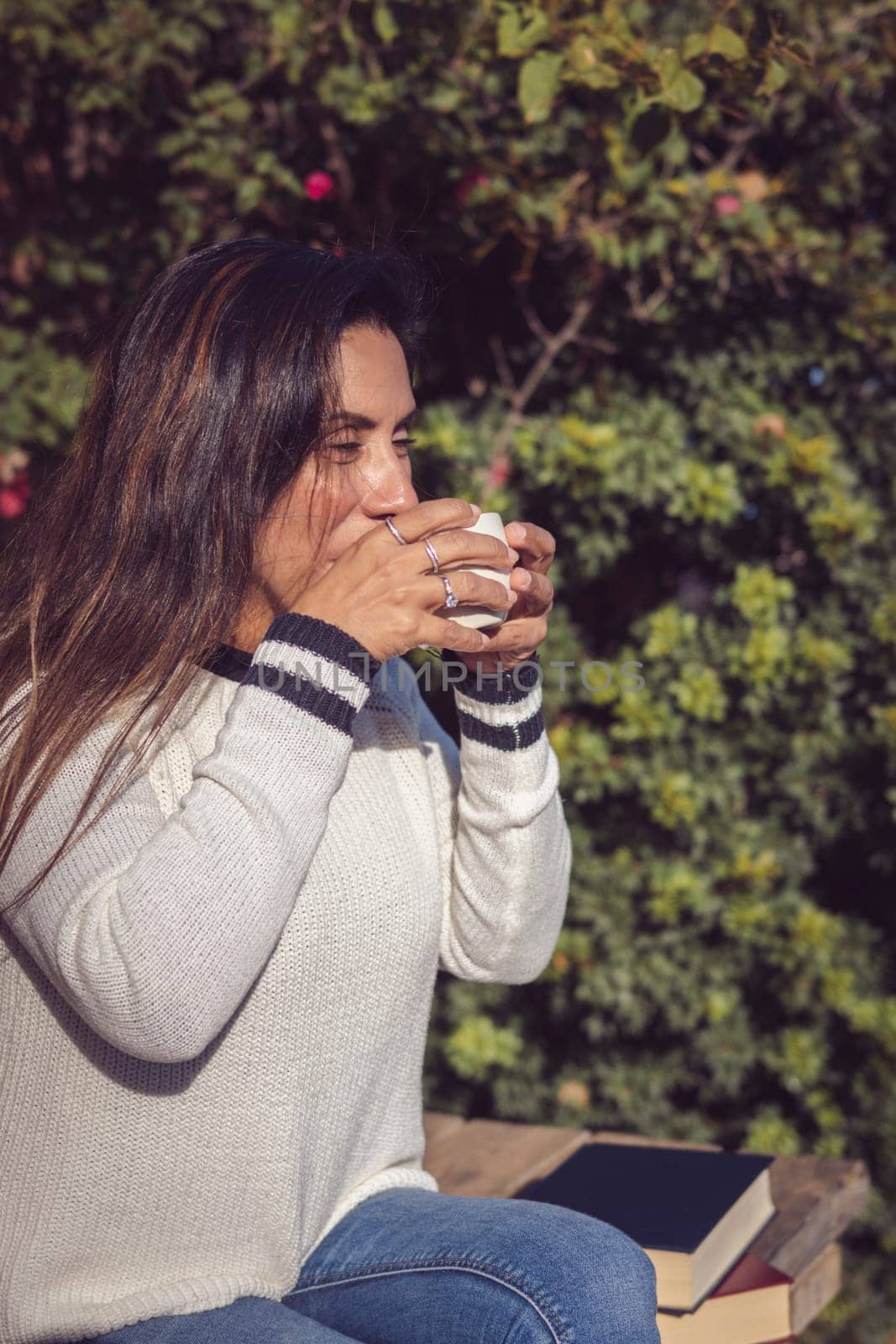 A Latina woman sits on a wooden bench in the garden, taking a sip of coffee while enjoying a moment of calm and relaxation. The natural beauty of the garden is appreciated in the background, creating a harmonious and peaceful atmosphere.