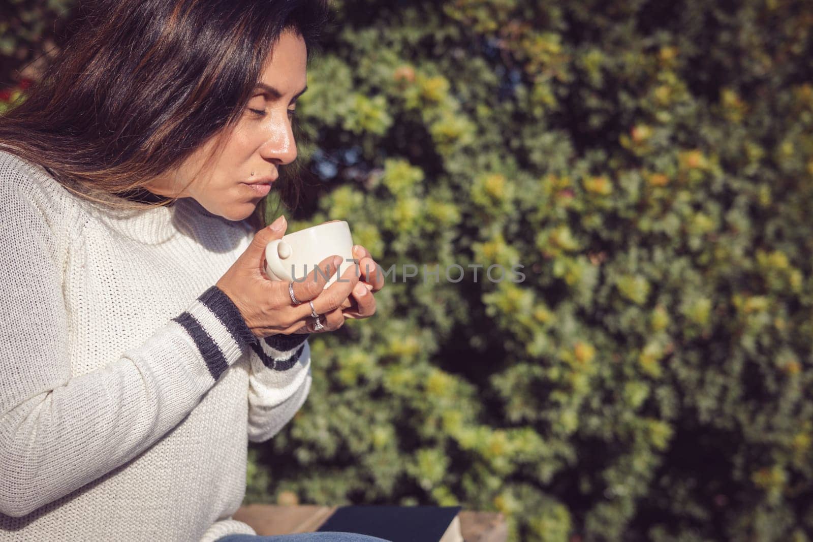 A Latina woman sits on a wooden bench in the garden, disconnecting from the outside world while taking a break to enjoy her coffee. The natural beauty of the garden conveys a sense of calm and peace, in harmony with the comforting sensation of drinking a hot beverage.