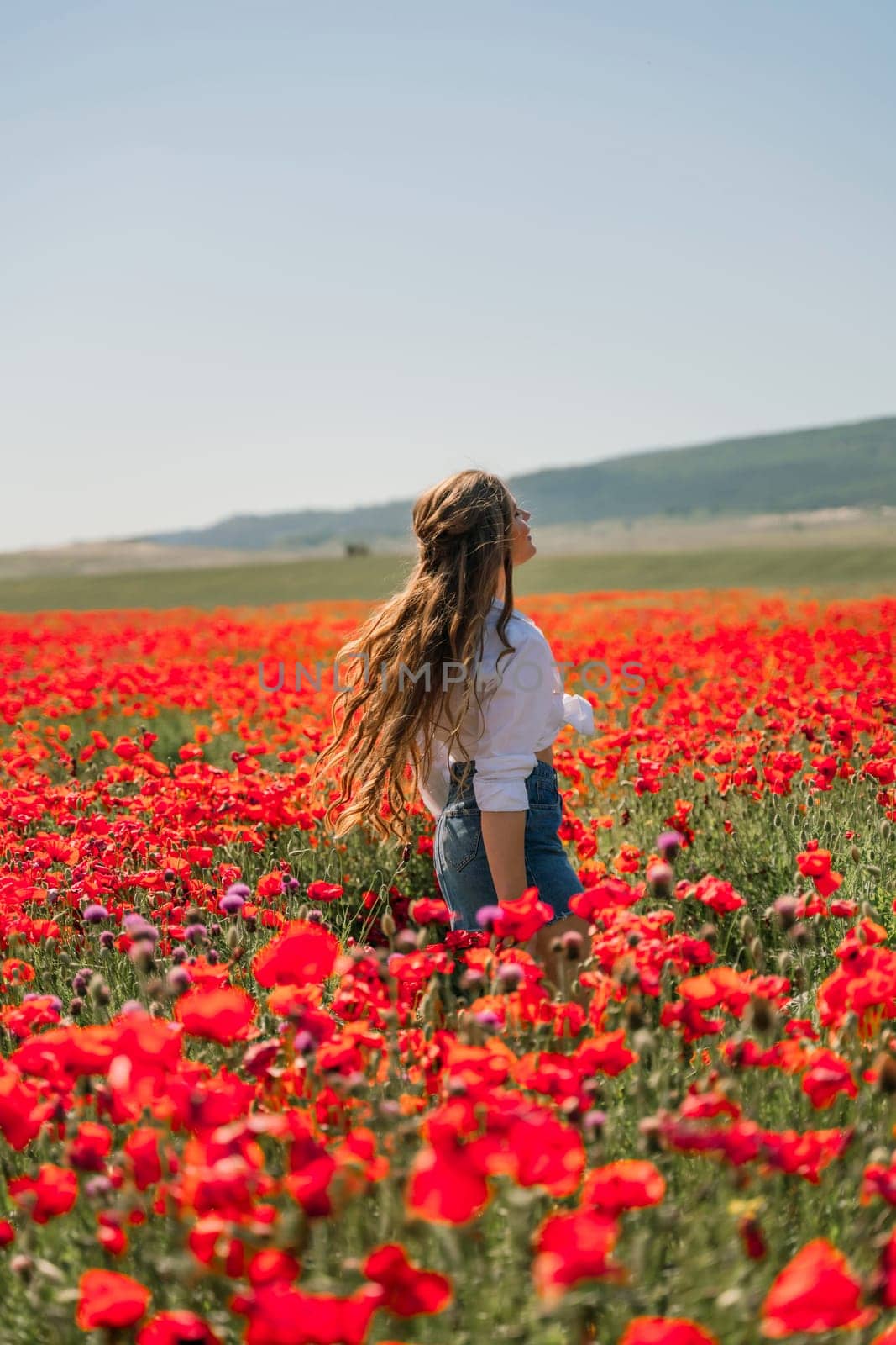 Happy woman in a poppy field in a white shirt and denim skirt with a wreath of poppies on her head posing and enjoying the poppy field. by Matiunina