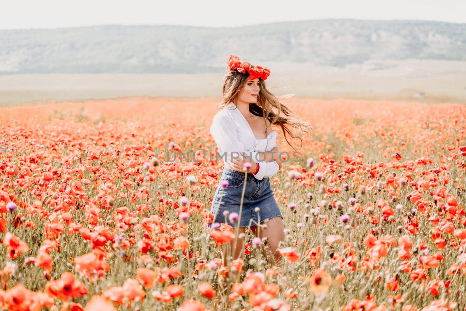 Happy woman in a poppy field in a white shirt and denim skirt with a wreath of poppies on her head posing and enjoying the poppy field