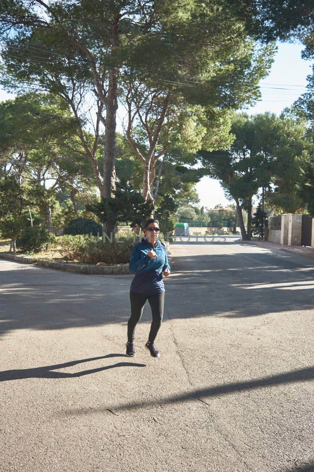 Mature fitness woman, between 30 and 50 years old, running, on the road, surrounded by pine trees, wearing sports glasses, bluetooth headphones, blue jacket, on a sunny winter day, Active Latin woman doing training, cardio workout, to burn calories