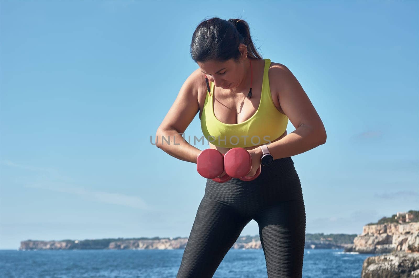 Middle-aged Latin woman, with yellow top, black leguin, with dumbbells, gymnastics, warmed up for training, burn calories, keep fit, outdoors by sea, blue sky, beautiful day, sunny winter, bluetooth headphones, smart watch, record the training