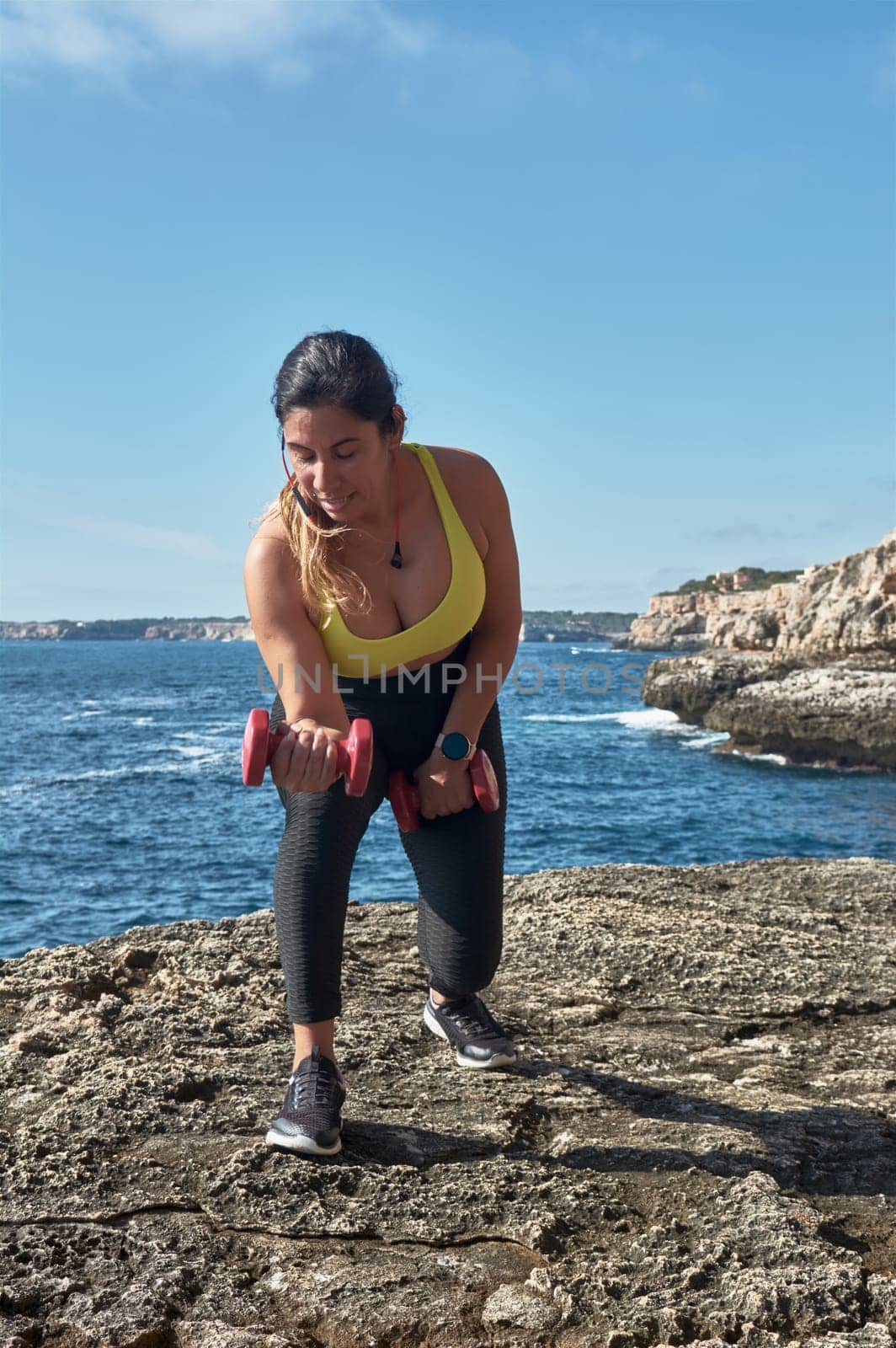Middle-aged Latin woman, with yellow top, black leguin, with dumbbells, gymnastics, warmed up for training, burn calories, keep fit, outdoors by sea, blue sky, beautiful day, sunny winter, bluetooth headphones, smart watch, record the training