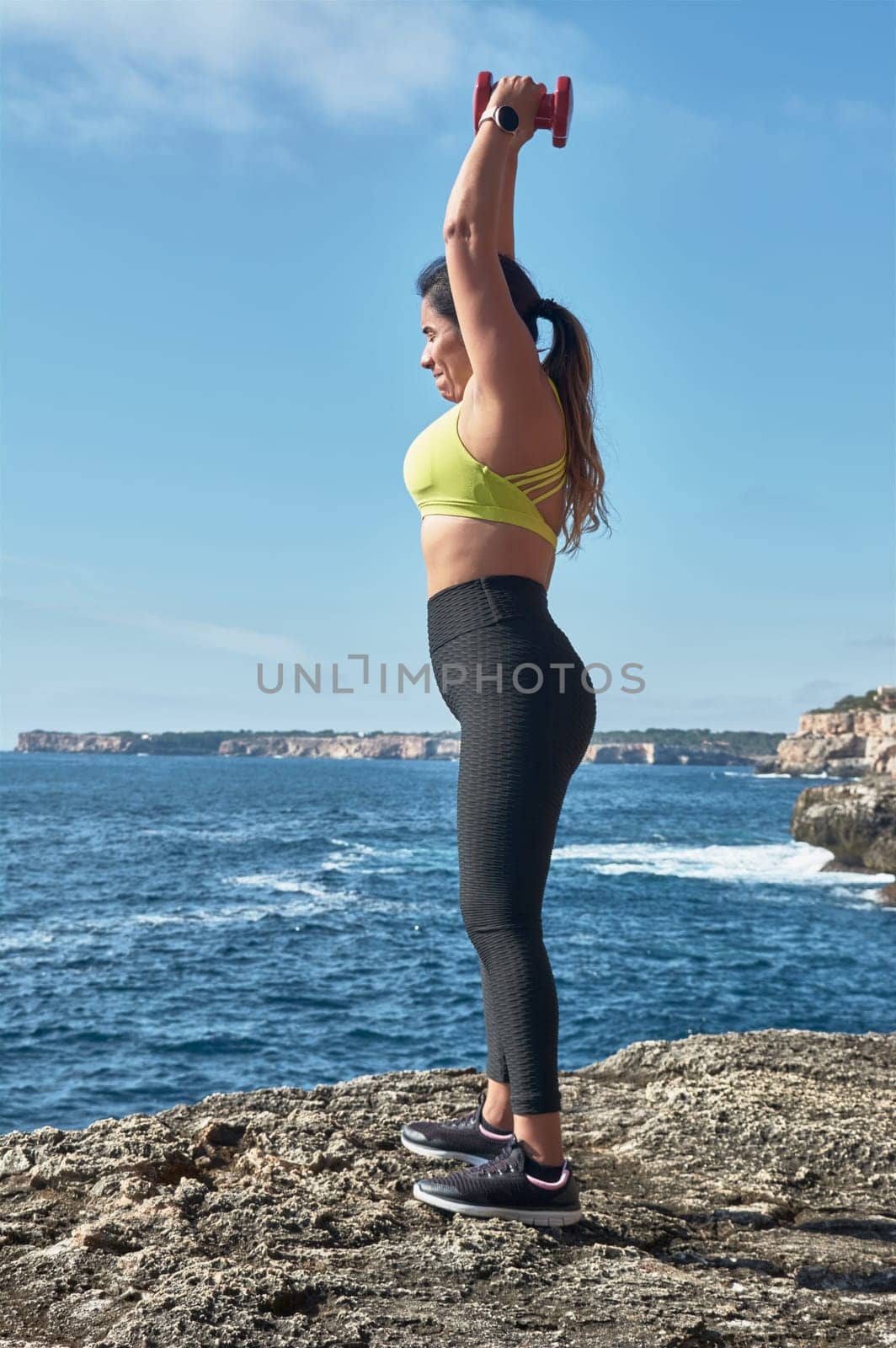 Middle-aged Latin woman, with yellow top, black leguin, with dumbbells, gymnastics, warmed up for training, burn calories, keep fit, outdoors by sea, blue sky, beautiful day, sunny winter, bluetooth headphones, smart watch, record the training