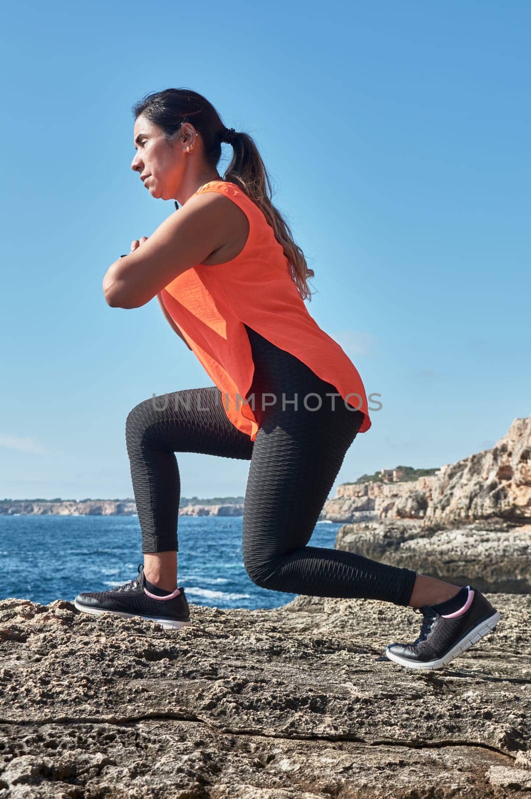 Latin woman, middle-aged, with orange top, black leggings, training, doing physical exercises, squats, burning calories, keeping fit, outdoors by the sea, blue sky, day, sunny winter, bluetooth headphones, smart watch, record the trainin