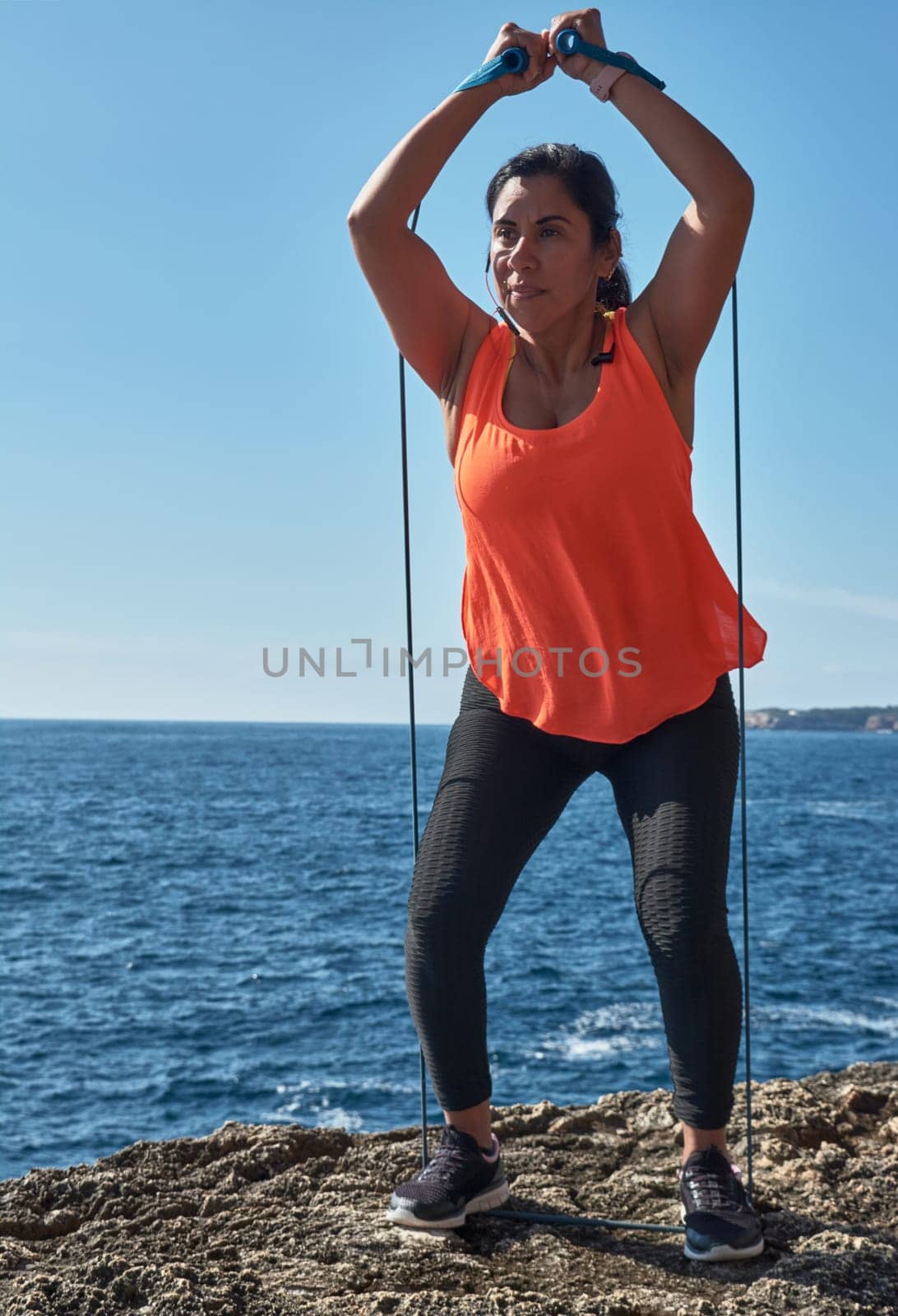 Latin woman, middle-aged, wearing sportswear, training, doing physical exercises, plank, sit-ups, climber's step, burning calories, keeping fit, outdoors by the sea, wearing headphones, smart watch