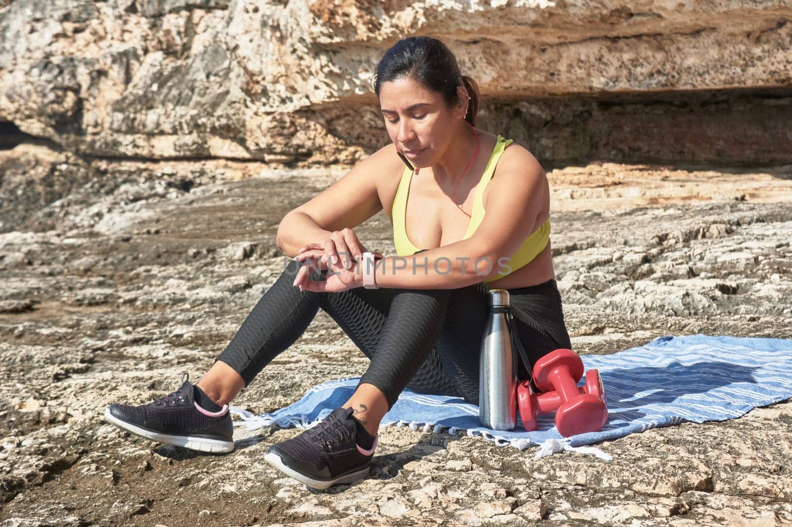 Latin woman, middle-aged, resting, regaining strength, eating, drinking water, after a gym session, burning calories, keeping fit, outdoors by the sea, wearing headphones and smart watch