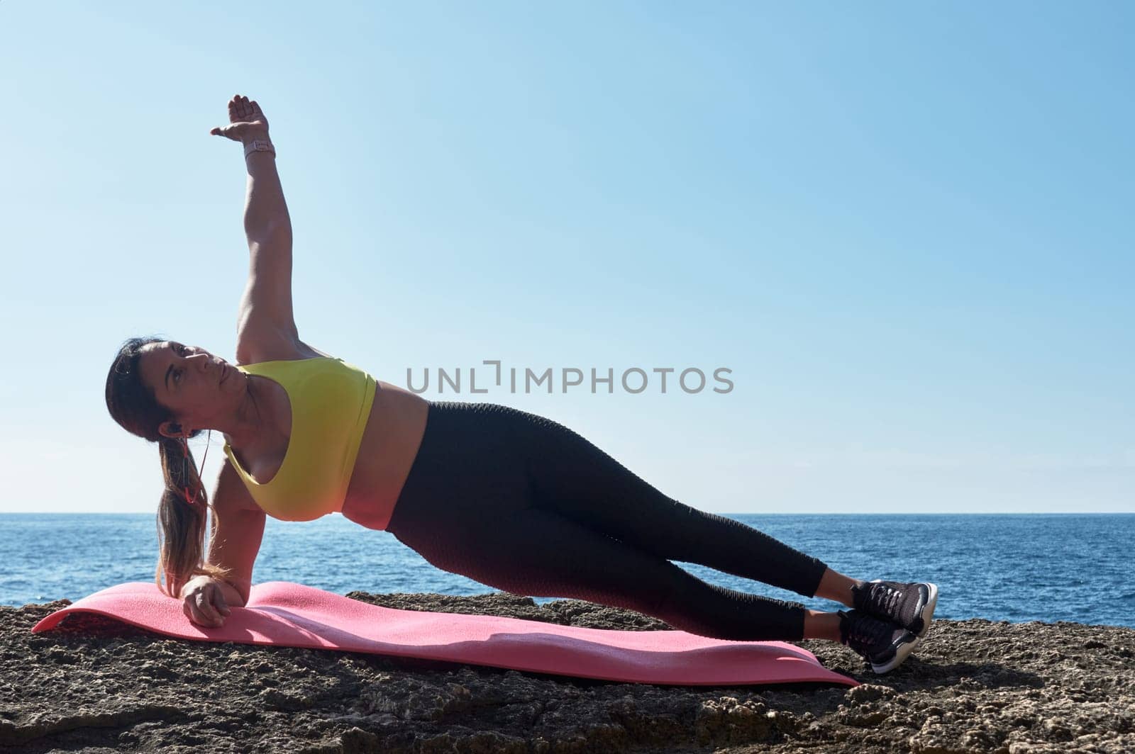 Latin woman, middle-aged, wearing sportswear, training, doing physical exercises, plank, sit-ups, climber's step, burning calories, keeping fit, outdoors by the sea, wearing headphones, smart watch