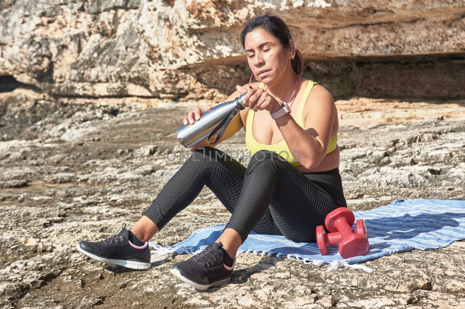 Latin woman, middle-aged, resting, regaining strength, eating, drinking water, after a gym session, burning calories, keeping fit, outdoors by the sea, wearing headphones and smart watch