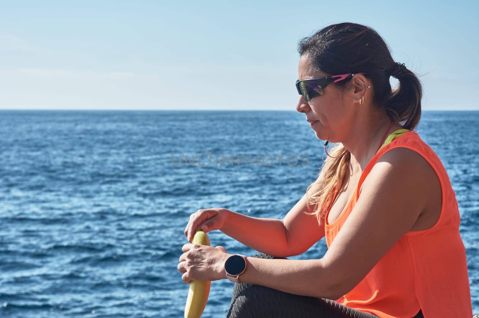 Latin woman, middle-aged, resting, regaining strength, eating, drinking water, after a gym session, burning calories, keeping fit, outdoors by the sea, wearing headphones and smart watch
