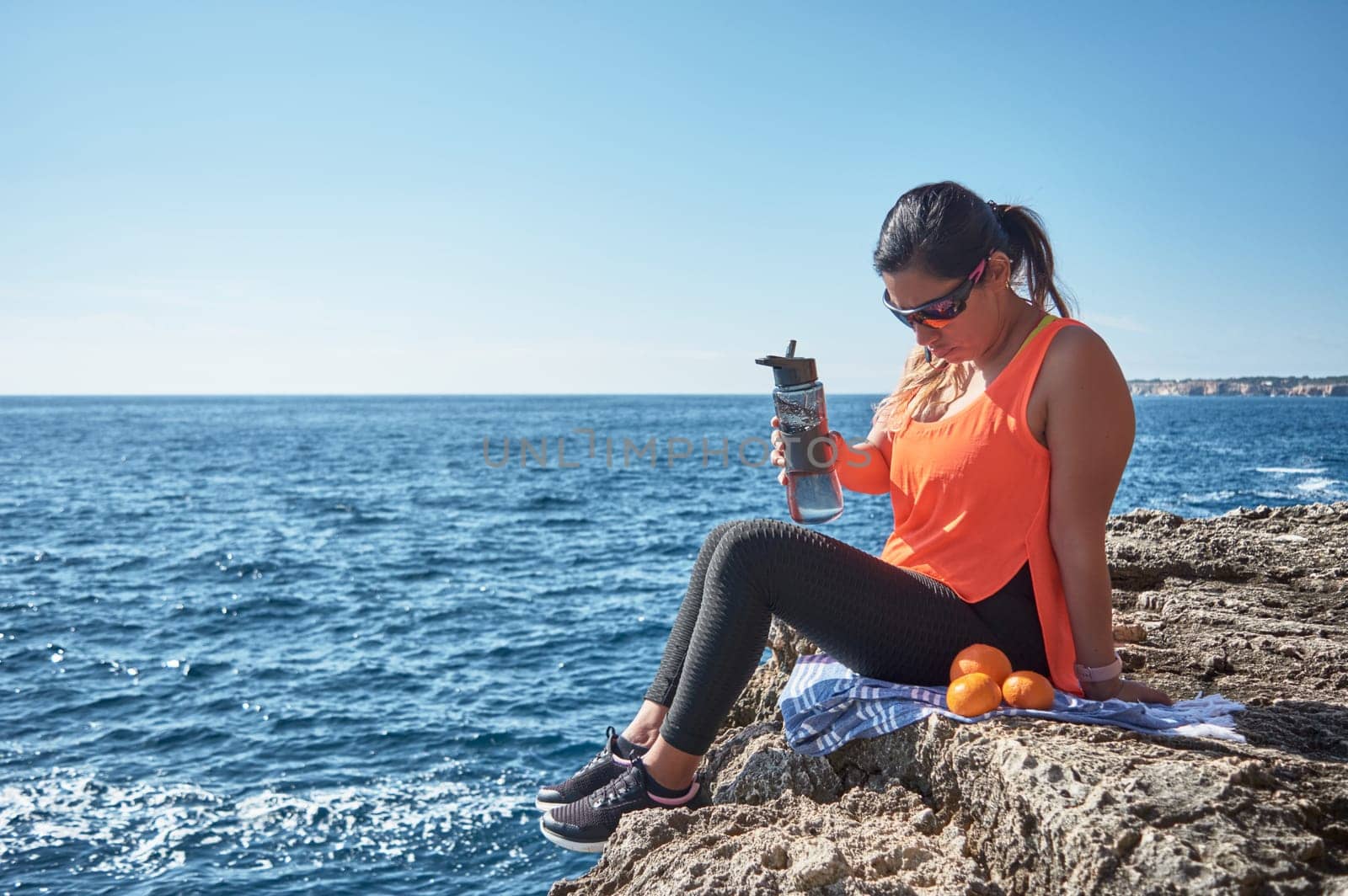 Latin woman, middle-aged, resting, regaining strength, eating, drinking water, after a gym session, burning calories, keeping fit, outdoors by the sea, wearing headphones and smart watch