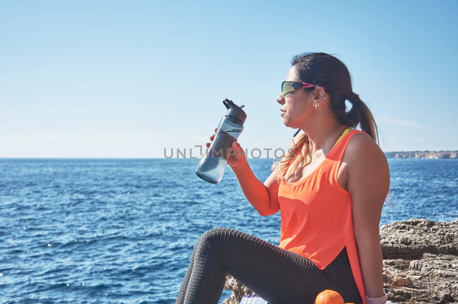 Latin woman, middle-aged, resting, regaining strength, eating, drinking water, after a gym session, burning calories, keeping fit, outdoors by the sea, wearing headphones and smart watch