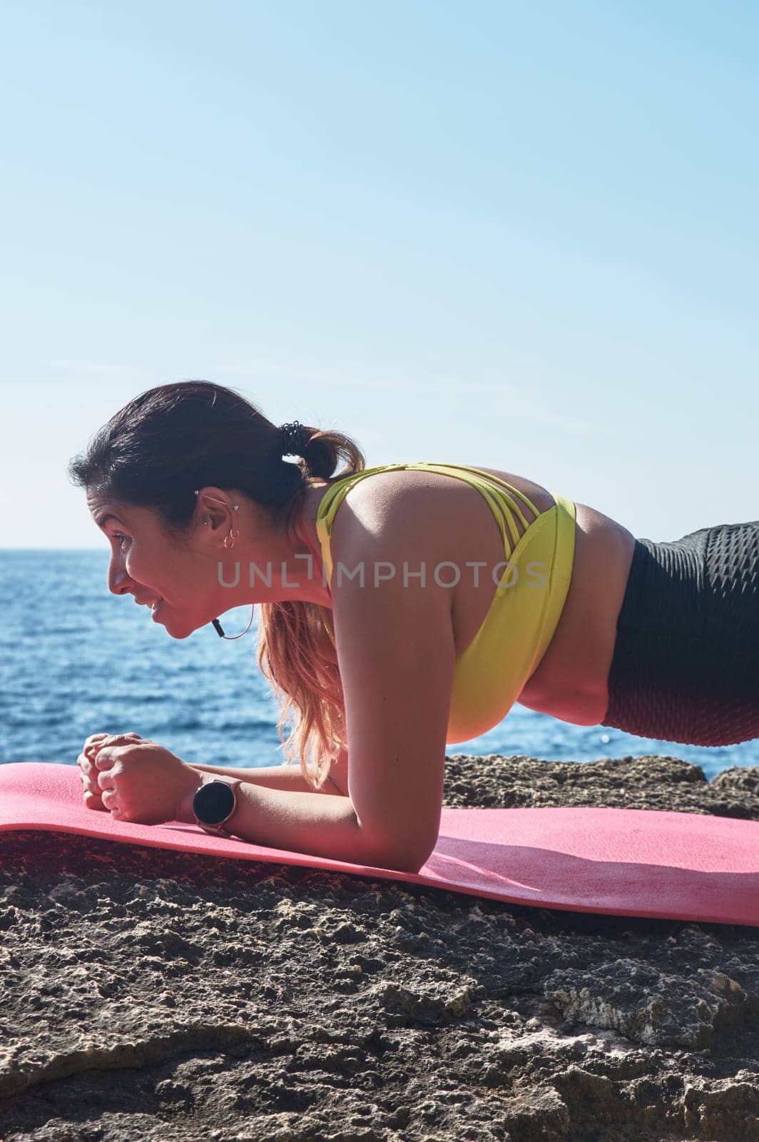 Latin woman, middle-aged, wearing sportswear, training, doing physical exercises, plank, sit-ups, climber's step, burning calories, keeping fit, outdoors by the sea, wearing headphones, smart watch