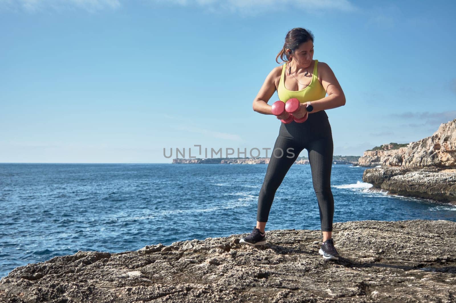 FITNESS WOMAN IN SPORTS SET TRAINING WITH ELASTIC BAND, WEIGHTS, GYM EXERCISES, IN FRONT OF THE WATER.