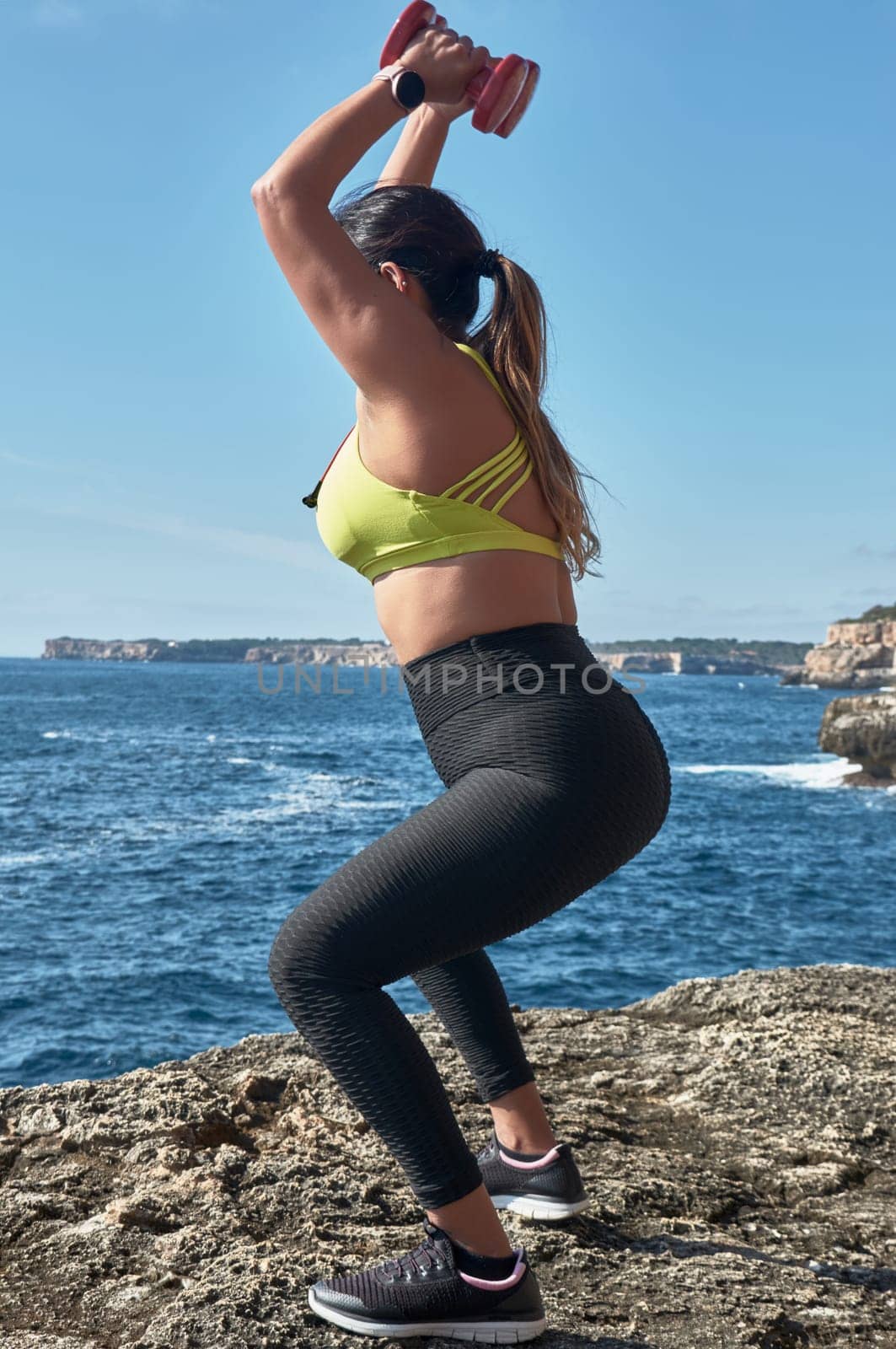 Latin woman, middle-aged, resting, regaining strength, eating, drinking water, after a gym session, burning calories, keeping fit, outdoors by the sea, wearing headphones and smart watch