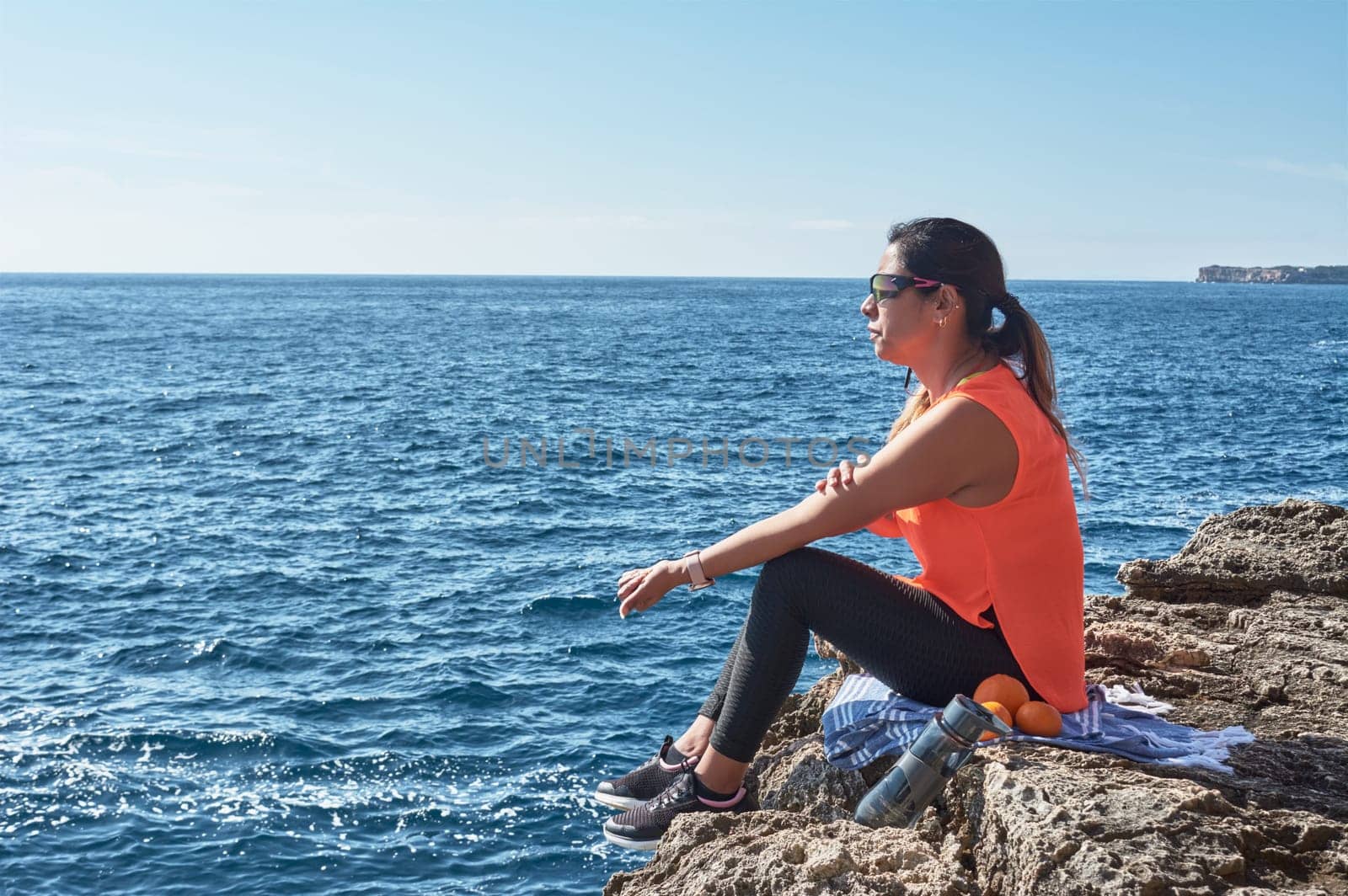 Latin woman, middle-aged, resting, regaining strength, eating, drinking water, after a gym session, burning calories, keeping fit, outdoors by the sea, wearing headphones and smart watch
