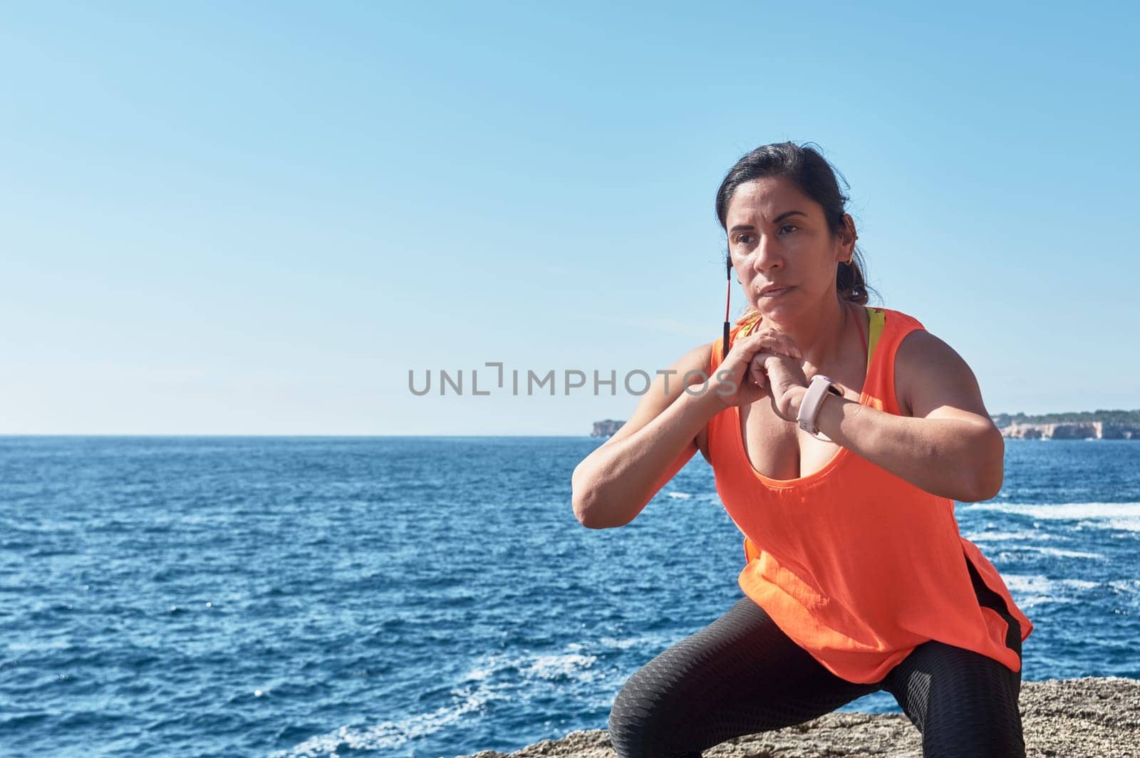 Latin woman, middle-aged, wearing sportswear, training, doing physical exercises, plank, sit-ups, climber's step, burning calories, keeping fit, outdoors by the sea, wearing headphones, smart watch,