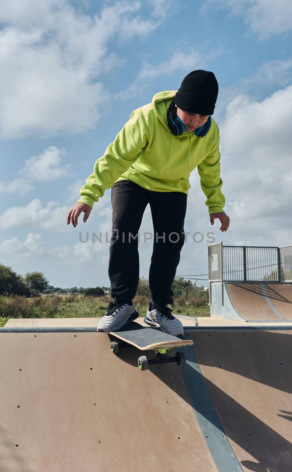 young, teenager, with a skateboard, diving down the slope, on a rink, skateboarding, wearing headphones, green sweatshirt, black hat, swinging, on a sunny day