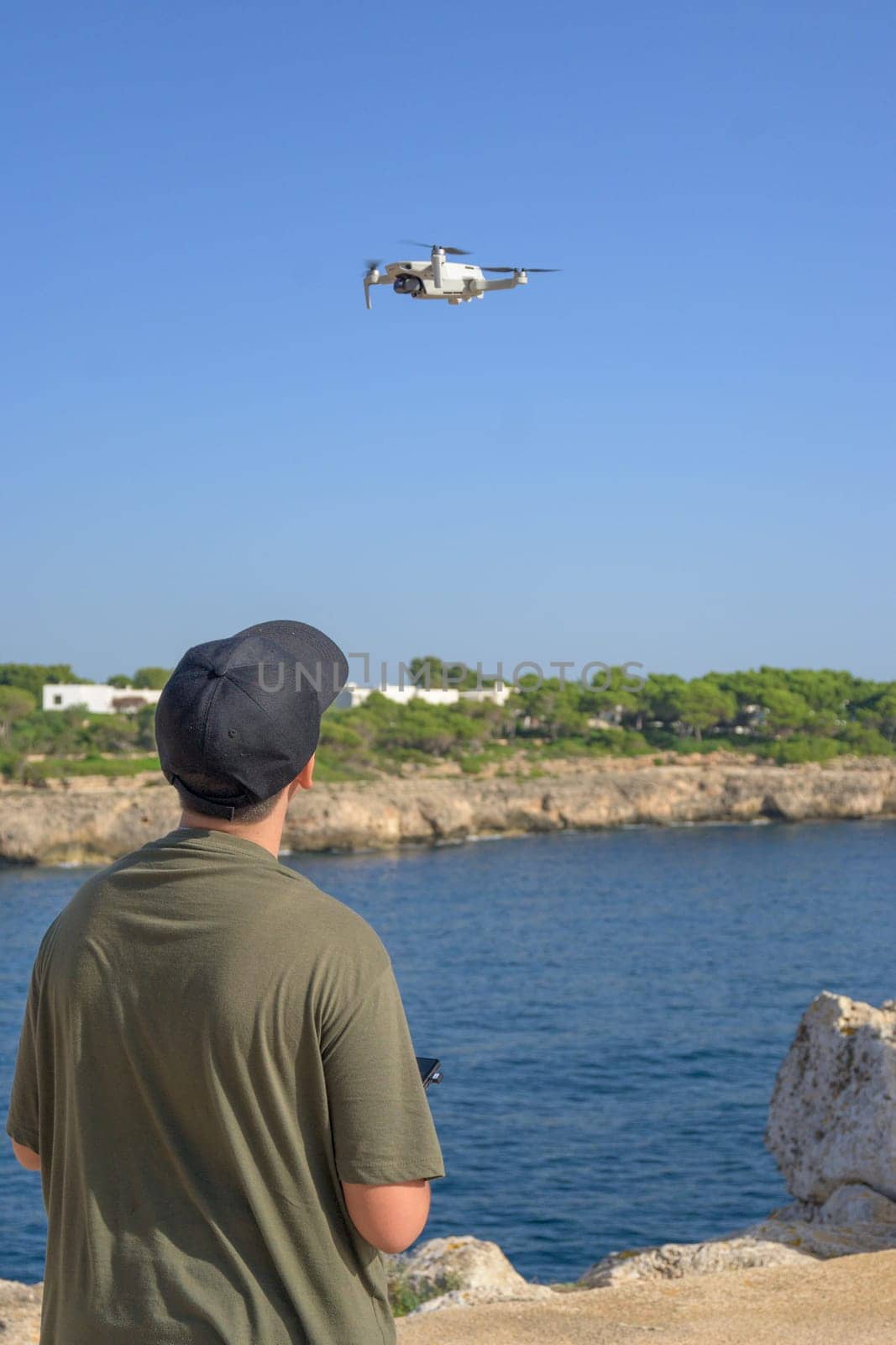 Rear view of teenage boy, flying drone on Mediterranean coast, against blue sky during sunny day Spain, Balearic Islands