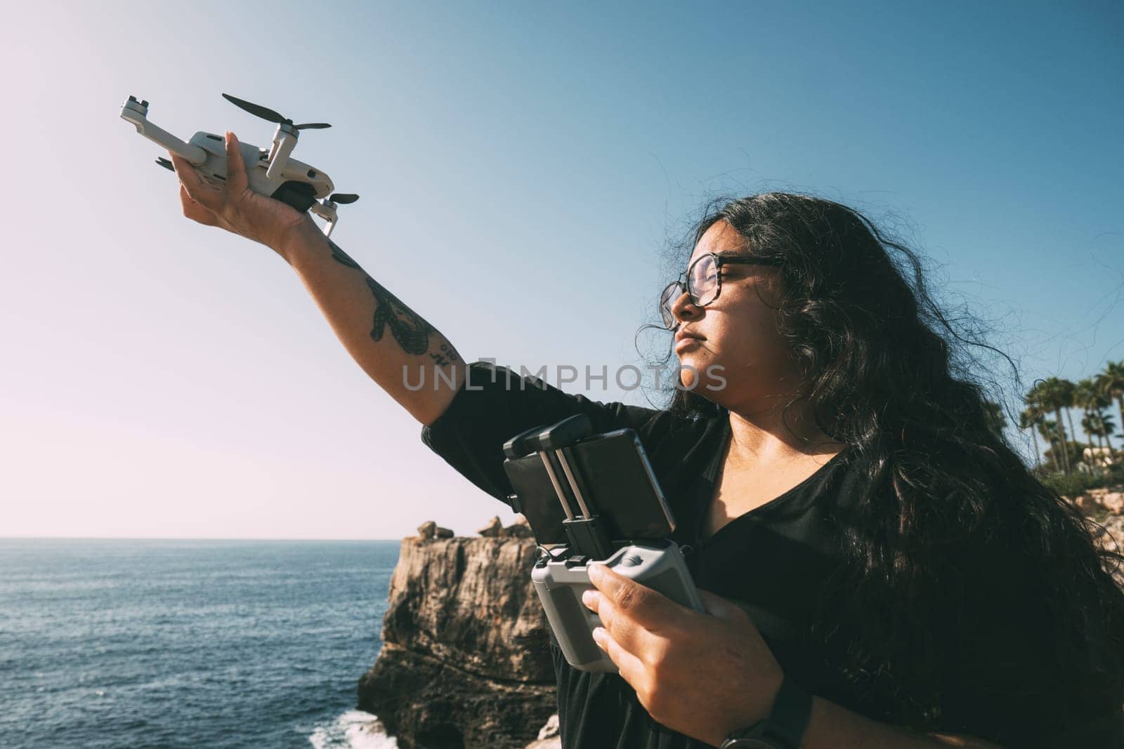 happy woman with drone, looking to the side black dress and remote control, on cliff, by the sea sunny day. technological concept,