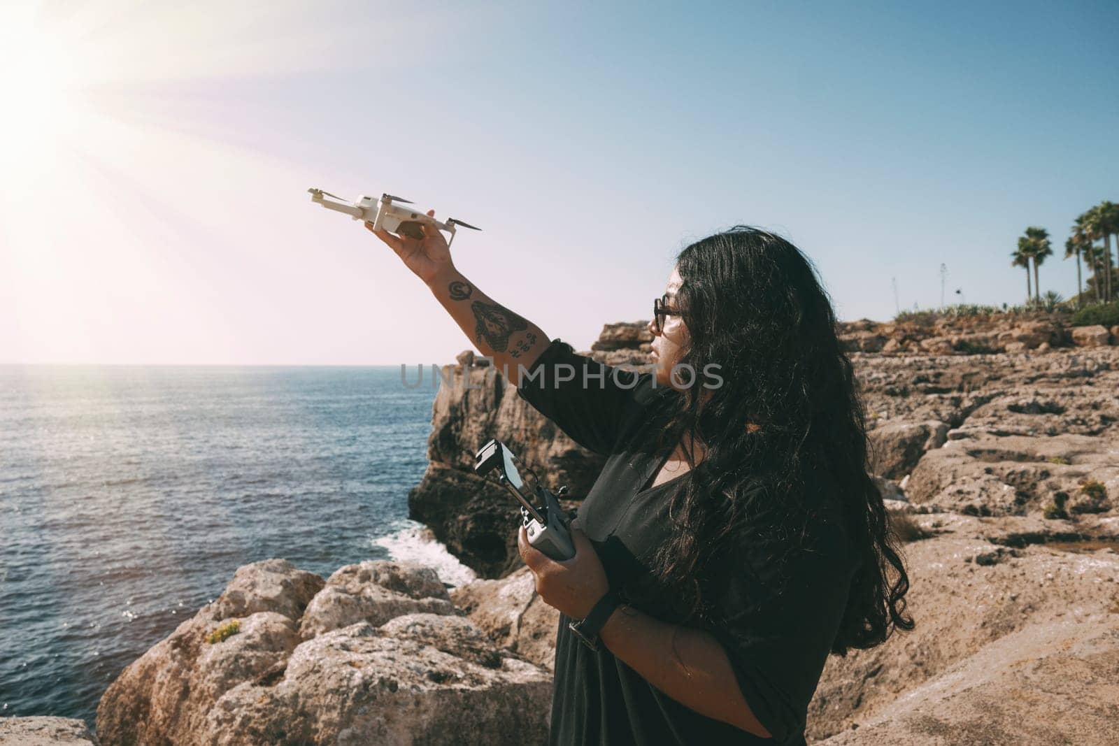happy woman with drone, looking to the side black dress and remote control, on cliff, by the sea sunny day. technological concept,