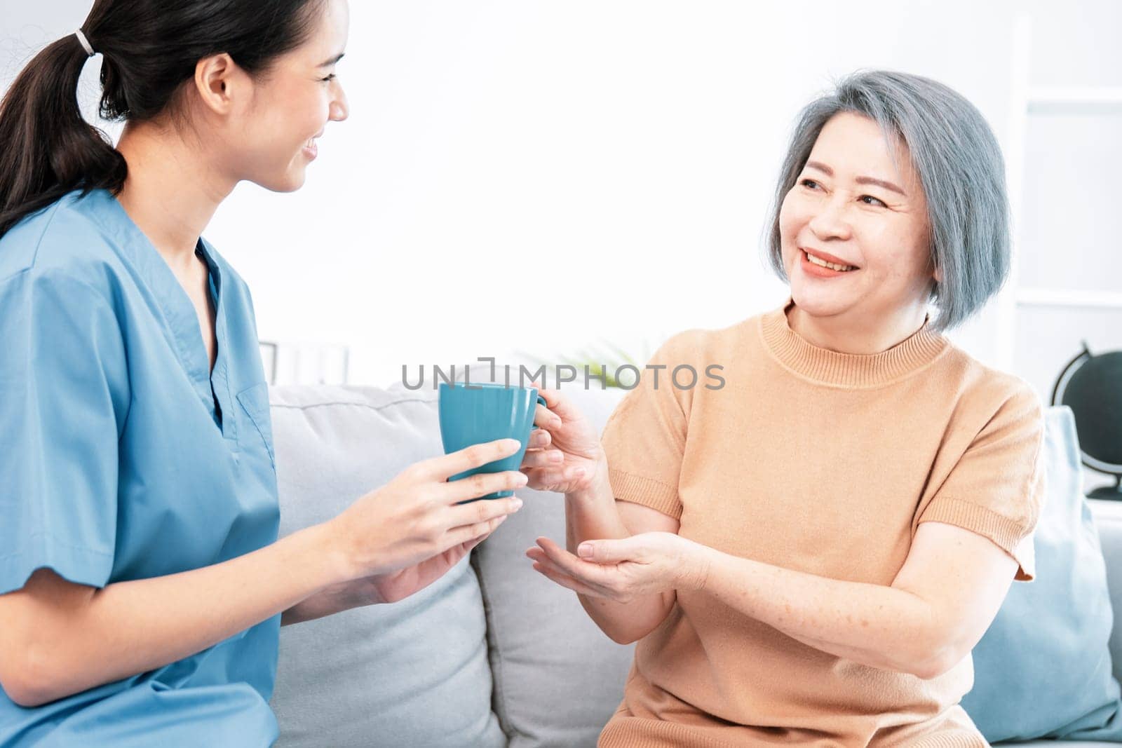 Female care taker serving her contented senior patient with a cup of coffee at home, smiling to each other. Medical care for pensioners, Home health care service.