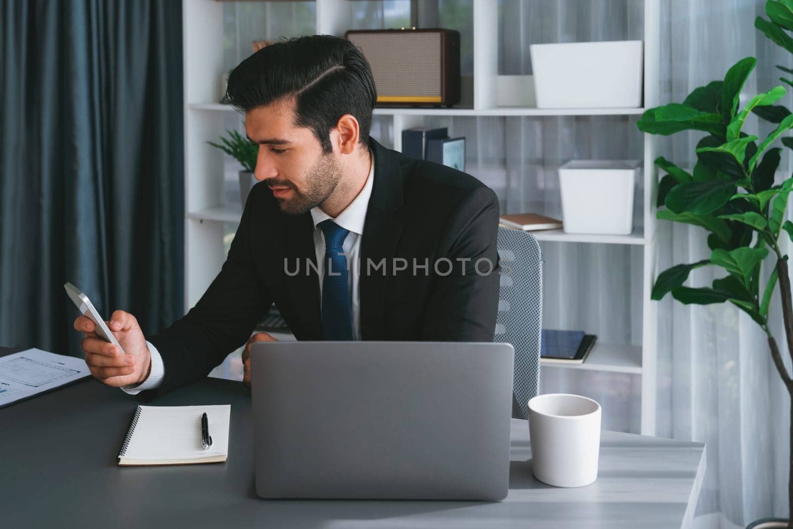 Diligent businessman busy talking on the phone call with clients while working with laptop in his office as concept of modern hardworking office worker lifestyle with mobile phone. Fervent