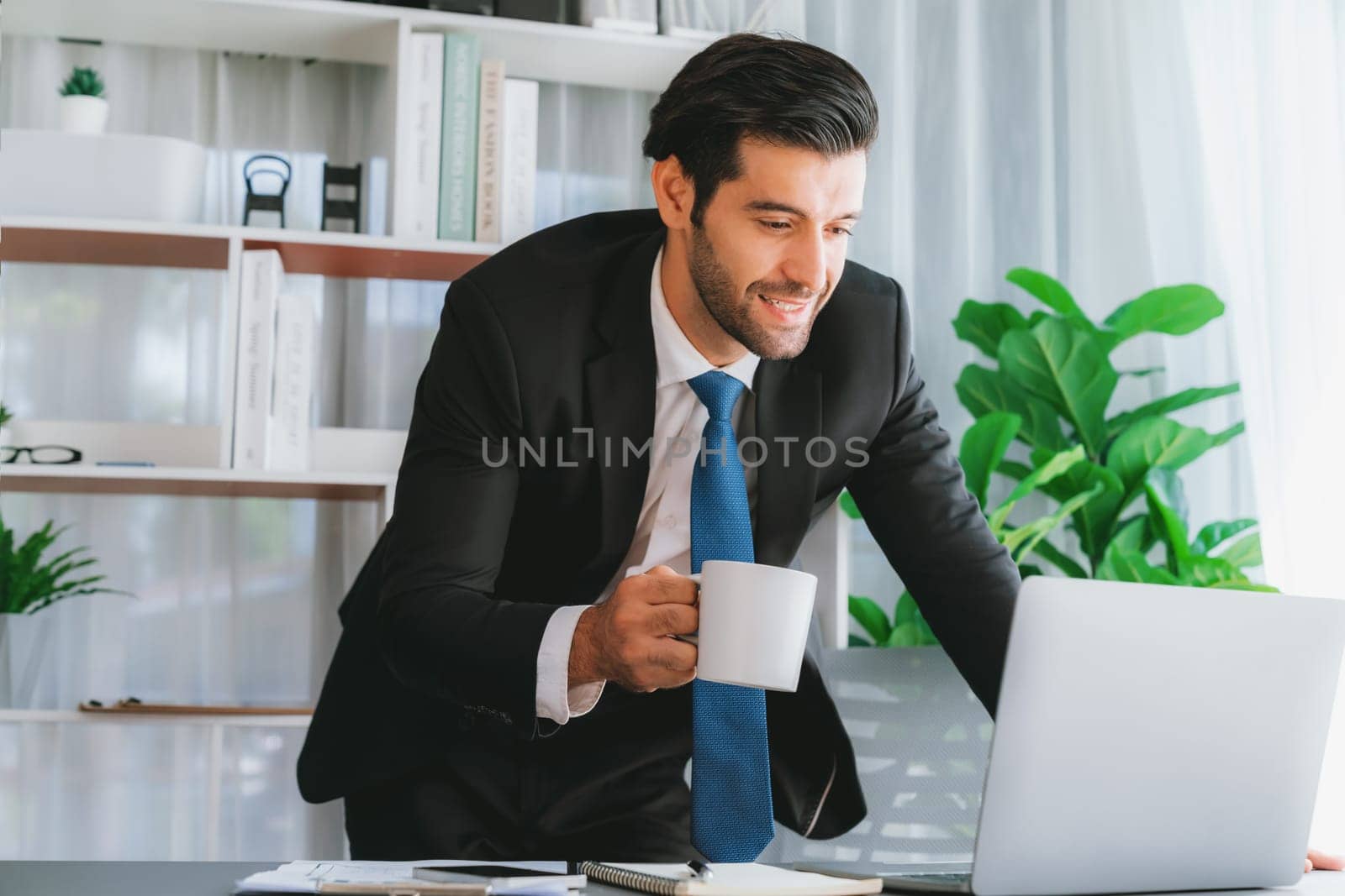 Modern professional businessman at modern office desk using laptop to work with coffee in his hand. Diligent office worker working on computer notebook in his office work space. fervent