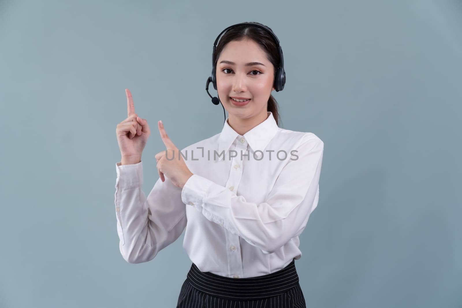 Asian female call center operator with smile face advertises job opportunity, wearing a formal suit and headset pointing finger for product on customizable isolated background. Enthusiastic