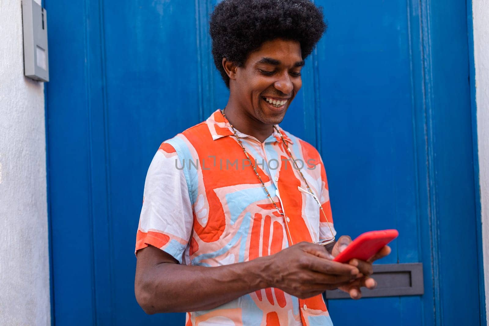 Happy black man with afro hairstyle using mobile phone to text a friend standing in the street. Blue door background. Copy space. Colourful image.