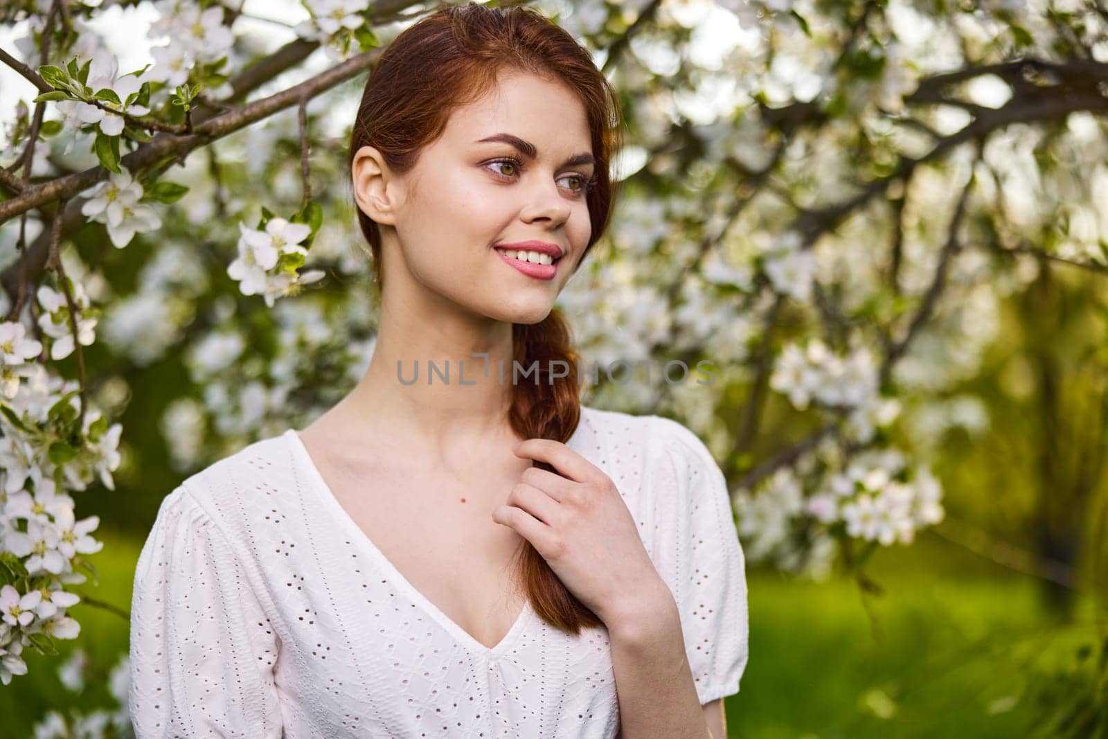 portrait of a joyful woman in a light dress against the background of a flowering tree, holding herself by her braid by Vichizh
