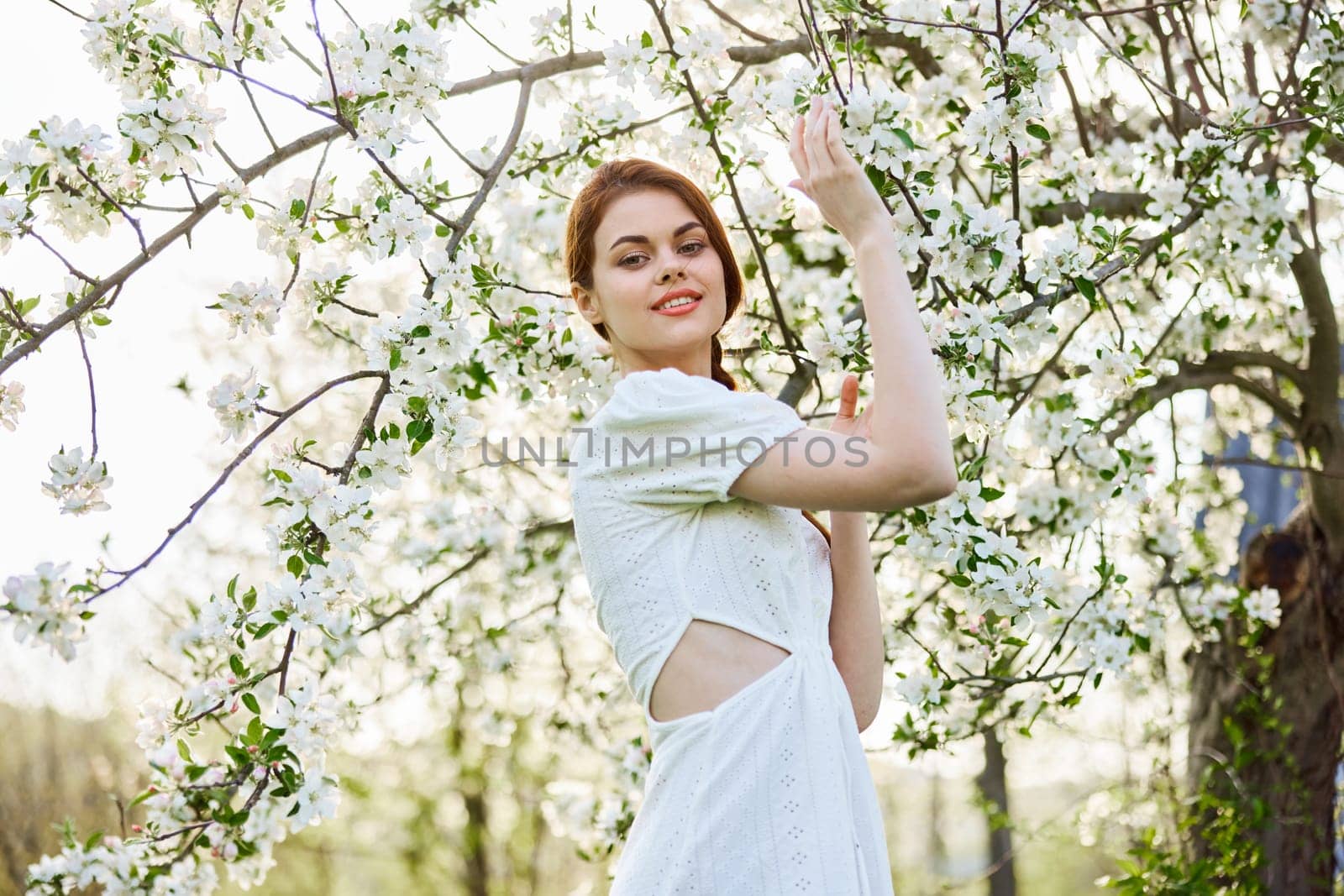 a slender, happy woman in a light dress poses next to a flowering tree in the countryside by Vichizh