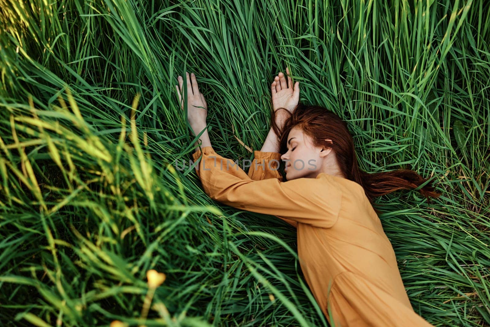 a close horizontal photo of a pleasant woman in a long orange dress resting lying in the tall grass with her eyes closed in sunny weather by Vichizh