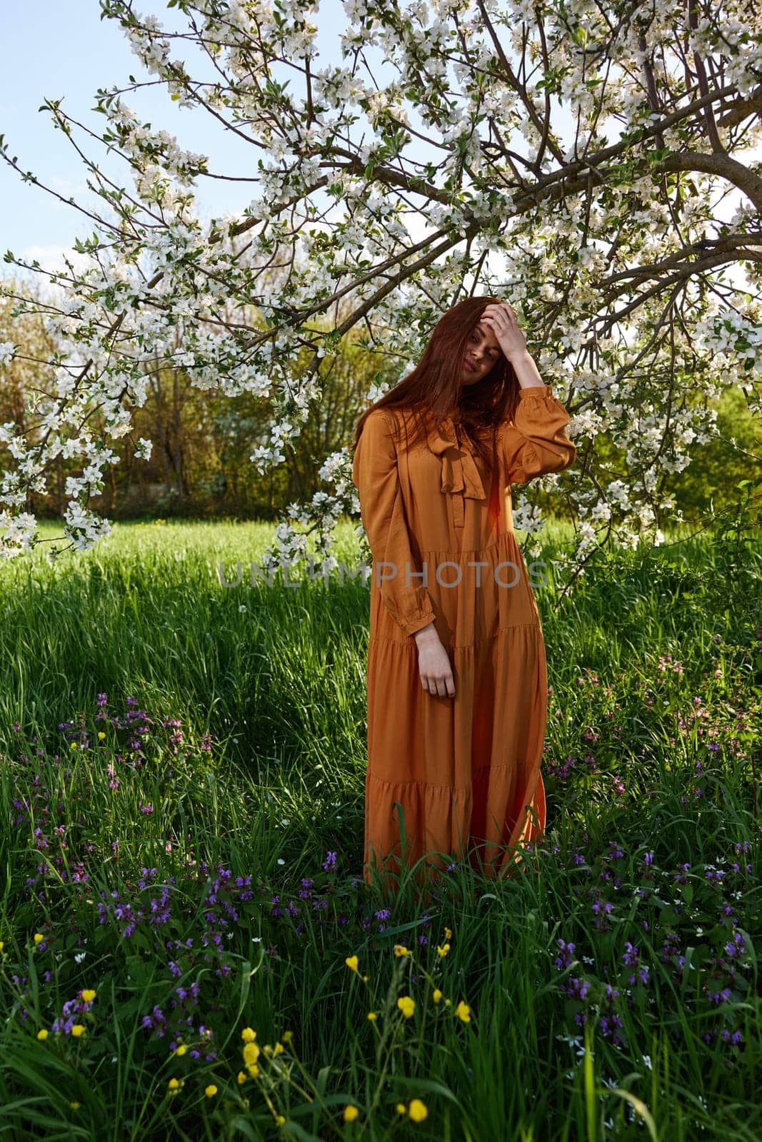 a happy, modest woman stands in an orange dress near a flowering tree and enjoys nature and a sunny summer day. High quality photo