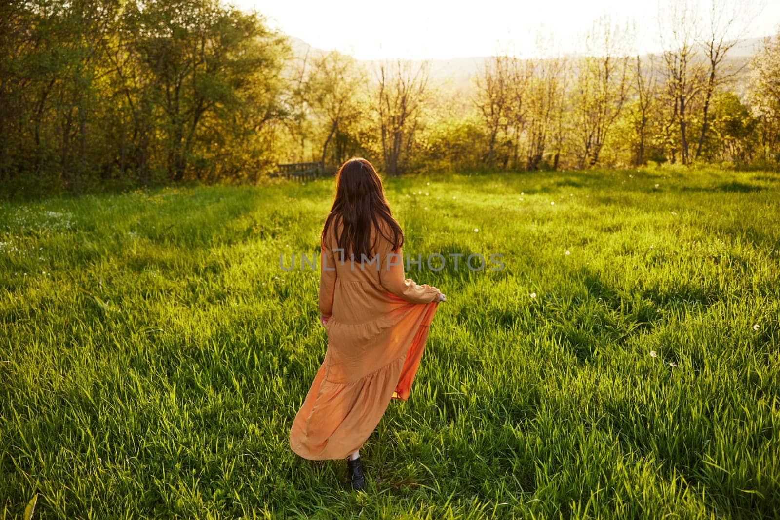 an attractive, slender, red-haired woman walks through a field during sunset, in a long orange dress enjoying unity with nature and relaxation holding the edge of her clothes by Vichizh