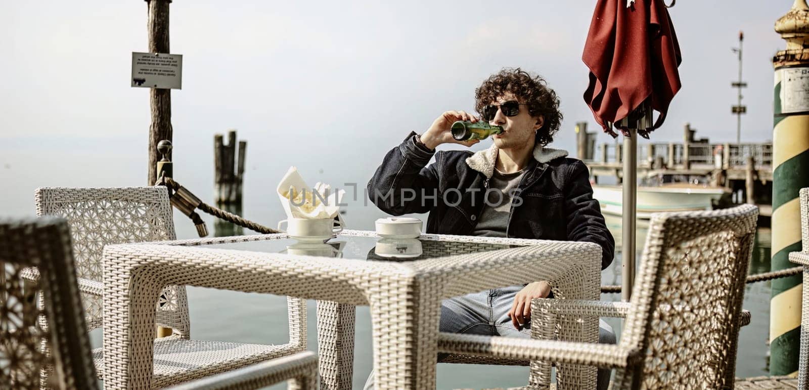 Melancholic Young Man Drinking Beer at Bar Table on Pier by pippocarlot