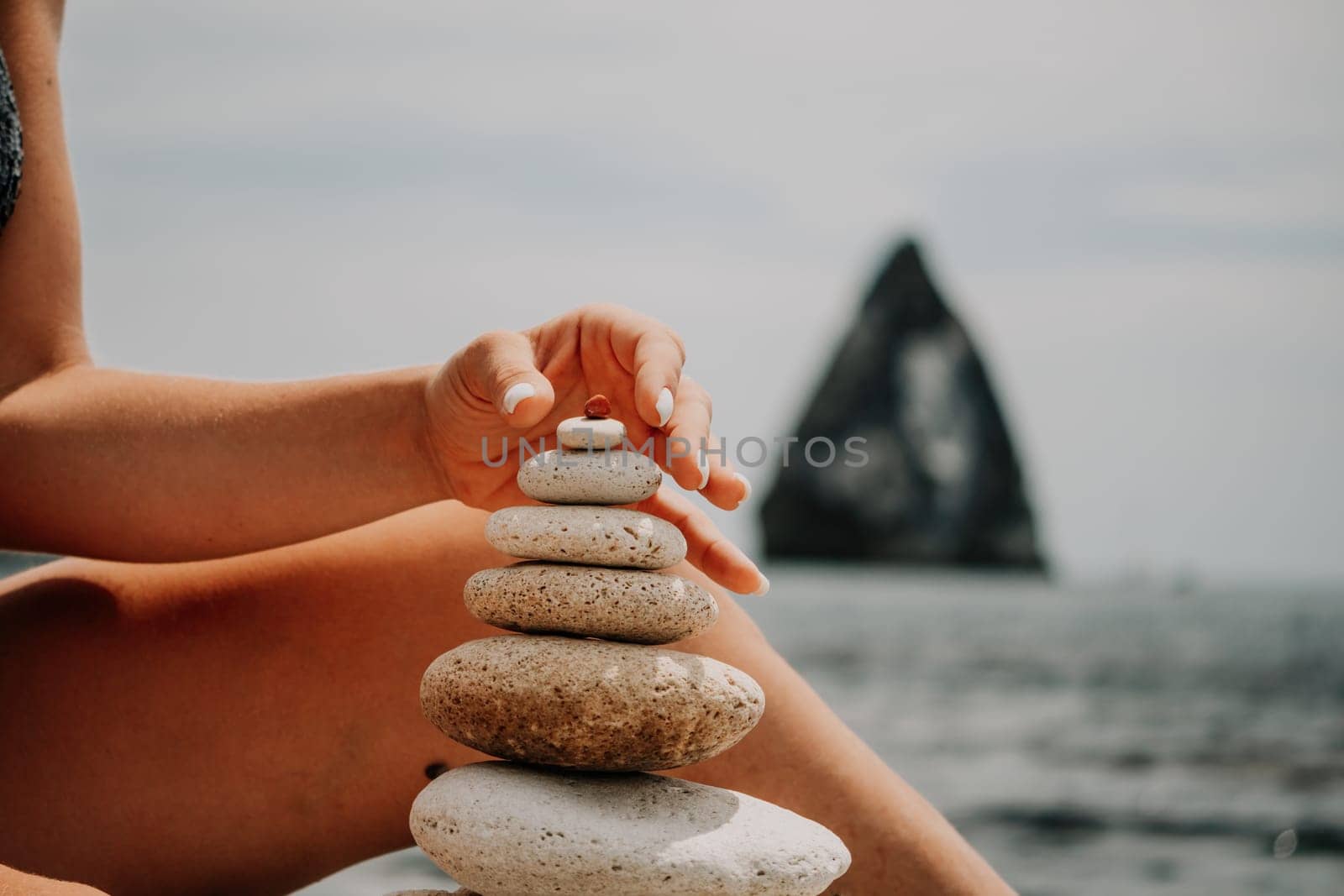 Woman building a stone pyramid on the seashore on a sunny day. Blue sea background. Travel destination, relaxation and meditation concept. by panophotograph
