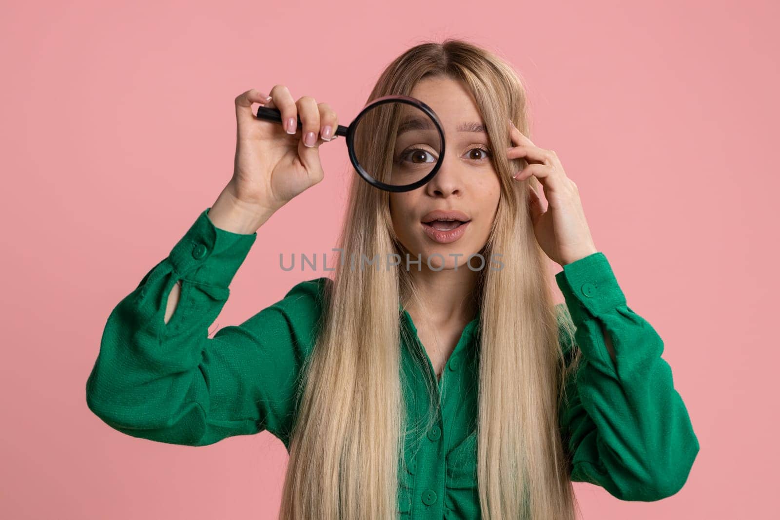 Investigator researcher scientist woman holding magnifying glass near face looking into camera with big zoomed funny eyes, searching analysing. Excited teen girl isolated on pink studio background