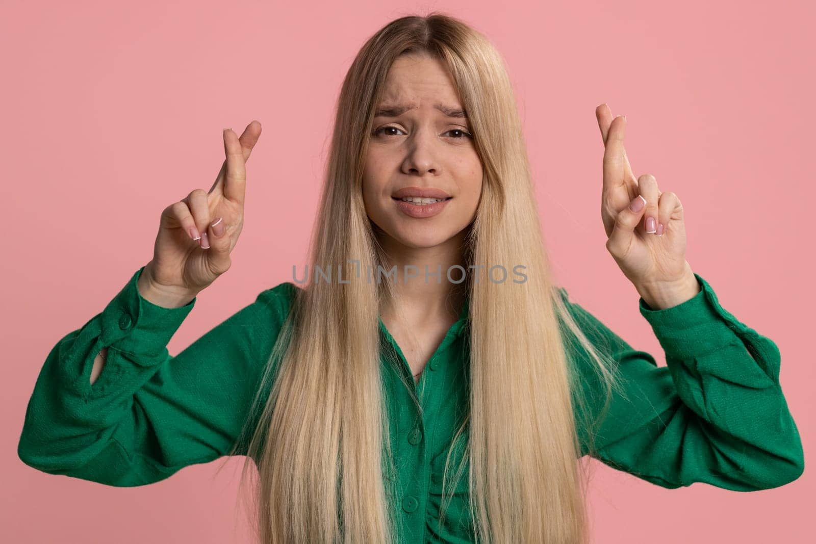 Young woman crossed fingers looking at camera asking for good luck news, wishing good education exam results dreaming about win victory in lottery jackpot. Teen girl isolated on pink studio background