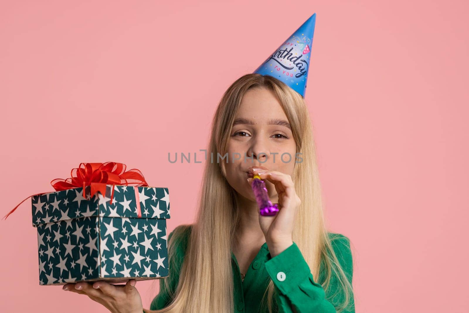 Happy young woman wearing birthday hat cone blow pipe celebrating success win victory. Excited lucky girl in anniversary party isolated on pink studio background indoors. People lifestyles concept