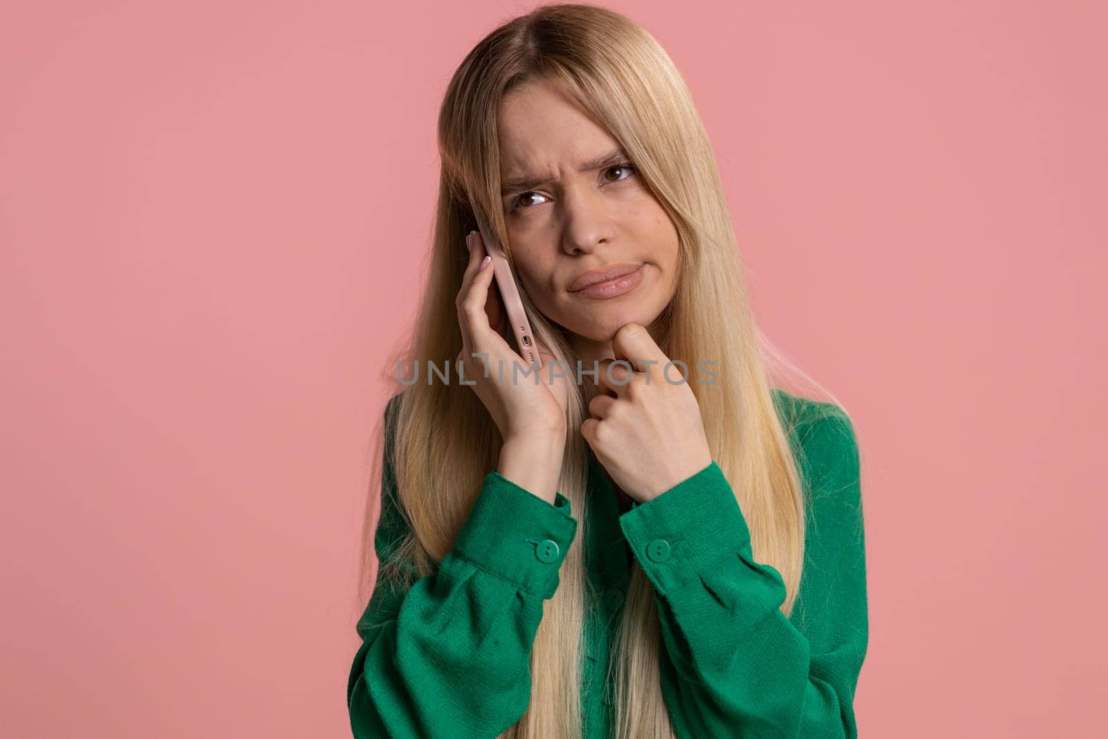 Disinterested young Caucasian woman having annoyed boring talk on smartphone. Tired sleepy adult girl talking on mobile phone with friend, making online conversation isolated on studio pink background