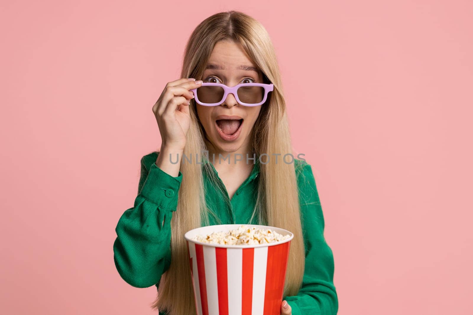 Excited pretty caucasian young woman in 3D glasses eating popcorn and watching interesting tv serial, sport game, film, online social media movie content. Teenager girl kid isolated on pink background